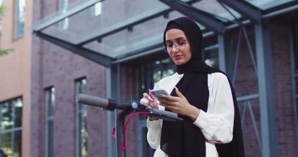 Piano medio colpo di giovani arabe belle donne in tradizionale velo noleggio scooter elettrico utilizzando l'app per cellulare. Belle femmine musulmane in hijab e occhiali in città. — Video Stock