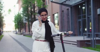 Geleneksel türbanlı neşeli Müslüman kadın portresi güzel şehir binasının yakınında kişisel e-scooterıyla sokakta yürüyor ve akıllı telefondan konuşuyor. Şehirde tesettürlü güzel Arap kadınlar.