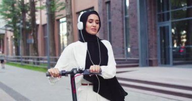 Geleneksel türbanlı neşeli Müslüman kadın portresi güzel bir şehir binasının yakınında kişisel e-scooter 'ıyla sokakta yürüyor ve kulaklıkla müzik dinliyor. Şehirde başörtüsü içinde güzel bir kadın..