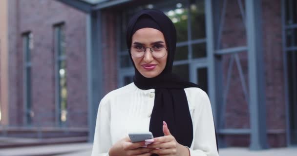 Close up of young Arabic beautiful woman in traditional headscarf and glasses lookingon phone and into camera. Close up of pretty cheerful muslim female in hijab at street. Islamic charming happy lady — Stock Video
