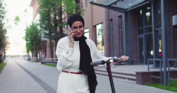 Porträt einer fröhlichen muslimischen Frau mit traditionellen Kopftüchern, die mit ihrem persönlichen E-Roller in der Nähe eines schönen städtischen Gebäudes auf der Straße unterwegs ist und mit dem Smartphone spricht. Hübsche arabische Frauen in Hijabs in der Stadt. — Stockvideo