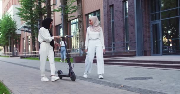 Plano medio de tiro de dos mujeres árabes hermosas en pañuelos tradicionales hablando, caminando por la calle con e-scooter personal cerca de hermoso edificio urbano. Bonitas hembras árabes en hiyabs en la ciudad. — Vídeo de stock