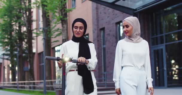 Plano medio de tiro de dos musulmanas hermosas mujeres en pañuelos tradicionales hablando, caminando por la calle con e-scooter personal cerca de hermoso edificio urbano. Bonitas hembras árabes en hiyabs en la ciudad. — Vídeos de Stock