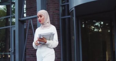 Üniversite bahçesinde elinde tablet ve kağıtlarla yürüyen kendinden emin Müslüman bir kadının portresi. Avrupa 'da okuyan genç bir göçmen kadın. Modern Orta Doğulu bayan yurtdışında eğitimin tadını çıkarıyor