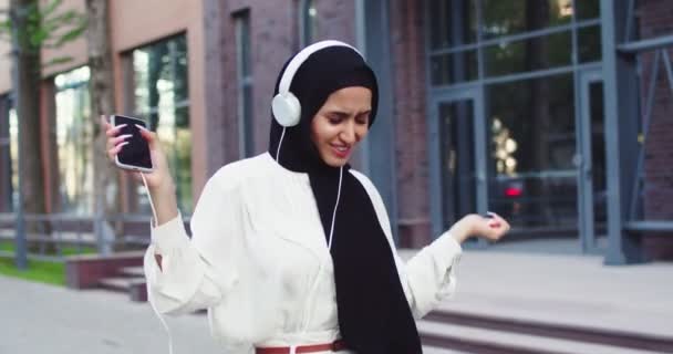 Moda düşkünü, neşeli müslüman, tesettürlü, cep telefonlu, şehir arka planında kulaklıkla müzik dinleyen genç bir kadın. Sokakta dans eden genç ve mutlu bir Arap kadının ağır çekimleri.. — Stok video