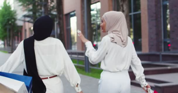 Middle plan back view shot of two young Arabic beautiful women in hijabs. Muslim females smiling, discussing, speaking, walking at street of a city, holding shopping bags in hands. — Stock Video
