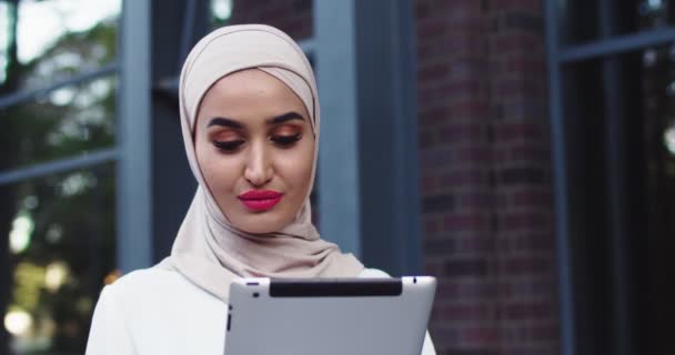 Close up portrait of young muslim pretty joyful woman in traditional hijab using tablet device at street. Urban landscape. Beautiful happy islamic female looking on gadget and at camera. Outdoors. — Stock Video