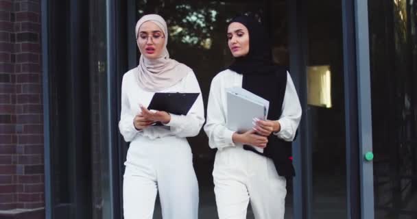 Piano di mezzo ripresa di due giovani arabe belle donne con il velo hijab, che camminano insieme giù per le scale con i documenti, parlando. Stile di vita moderno, tecnologia, discussioni commerciali — Video Stock