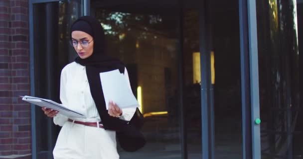 Portret pewnej siebie muzułmanki w okularach spacerującej po dziedzińcu uniwersyteckim z tabliczką i papierami. Młoda imigrantka studiująca w Europie. Nowoczesny Bliski Wschód pani korzystających edukacji za granicą — Wideo stockowe