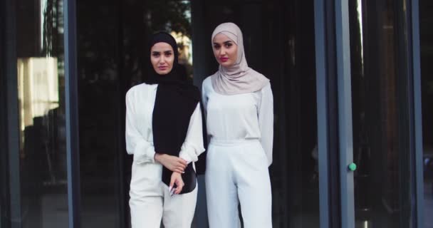 Middle plan shot of two young Arabic beautiful women in traditional headscarf and glasses looking at camera. Pretty cheerful muslim females in hijab at street. Islamic charming happy ladies — Stock Video