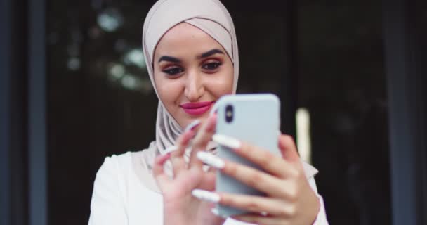 Arabisk kvinde med traditionelle tørklæder kigger på telefonen opholder sig på stree og glad smil. Temmelig muslimsk kvinde i hijabs ved hjælp af web-kamera på smartphone, hvilket gør selfie, swiping på skærmen. – Stock-video