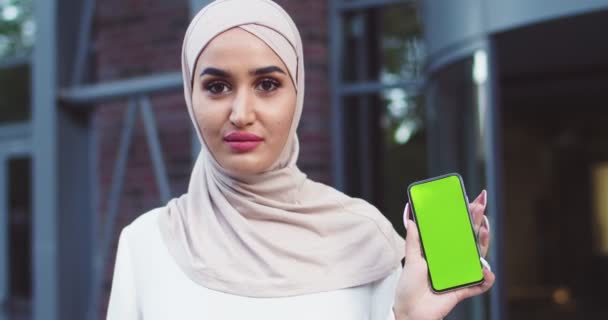 Close up de mulher muçulmana confiante em hijab preto segurando smartphone com greenscreen. Senhora bonita moderna em lenços de cabeça tradicionais brancos posando ao ar livre. Senhoras felizes islâmicas em roupa elegante. — Vídeo de Stock