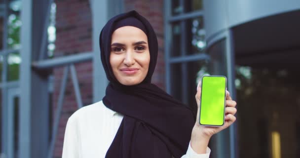 Close-up van zelfverzekerde moslimvrouw in zwarte hijab met smartphone met greenscreen. Moderne mooie dame in zwarte traditionele hoofddoeken poseren buiten. Islamitische vrolijke dames in stijlvolle outfit. — Stockvideo
