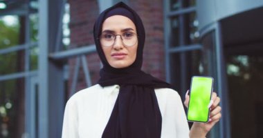 Siyah tesettürlü, gözlüklü, yeşil ekranlı akıllı telefon tutan kendine güvenen Müslüman bir kadına yaklaş. Geleneksel siyah eşarplı modern güzel bayan dışarıda poz veriyor. İslami mutlu bayanlar