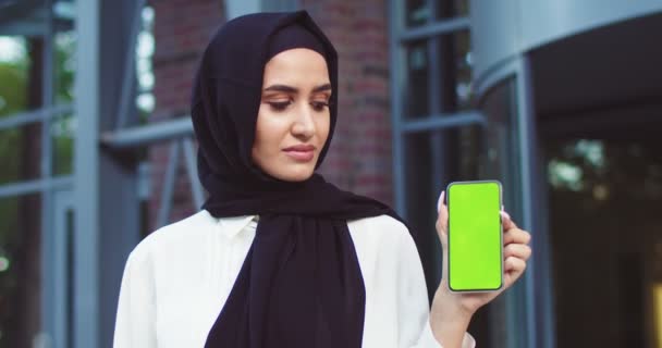 Close up de mulher muçulmana confiante em hijab preto segurando smartphone com greenscreen e olhando para ele, em seguida, para a câmera. Moderna bela senhora em preto tradicional lenços posando ao ar livre. — Vídeo de Stock