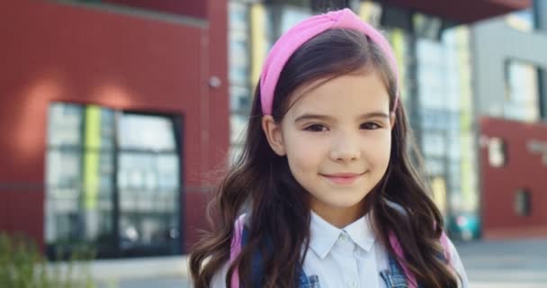 Nahaufnahme Porträt der bezaubernden schönen kaukasischen kleinen Mädchen lächelt vor der Schule. Ziemlich niedliches Schulkind, das mit einem Lächeln im Gesicht allein draußen steht. Zurück zur Schule. Schülerkonzept — Stockvideo