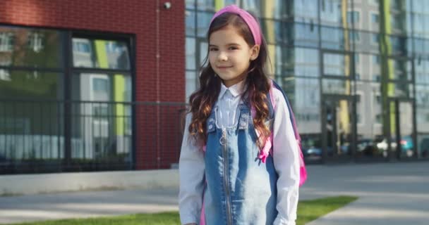 Retrato de niña linda linda linda feliz escuela en banda de pelo rosa sonriendo mientras está de pie en el patio de la escuela. Un niño muy agradable de buen humor al aire libre. Estudiante junior. Concepto educativo — Vídeos de Stock