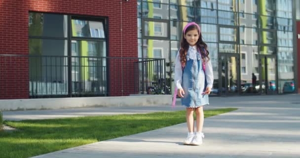 Vrolijk schattig mooi schoolmeisje in roze hoofdband glimlachend terwijl ze alleen op het schoolplein staat. Mooie blanke leerling in een goede bui buiten. Vrouwelijke junior student. Onderwijsconcept — Stockvideo