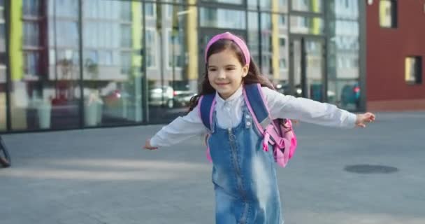 Ritratto di ragazza caucasica carina felice scuola con zaino rosa in esecuzione sul cortile della scuola dopo le lezioni. Bella gioiosa piccola studentessa junior che sorride all'aperto per strada. Concetto di emozioni — Video Stock