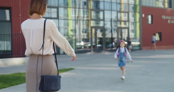 Posteriore della donna caucasica in piedi all'aperto e in attesa di figlia studente junior da scuola. Gioiosa scolaretta carina in fascia rosa che corre dalla madre dopo le lezioni. Concetto educativo — Video Stock