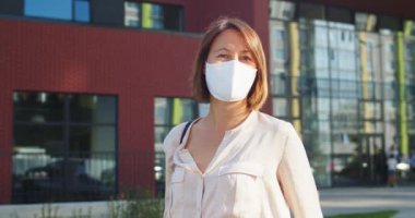 Karantinada güneşli bir günde okulun önünde maske takan mutlu güzel bayanı kapatın. Şehrin sokaklarında neşeli, beyaz, güzel bir kadın. Arka plandaki insanlar. Portre konsepti