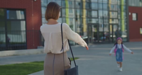 Vacker glad vit kvinna står utomhus och väntar på liten dotter från skolan. Glada söt tjej i rosa pannband kramar mamma efter lektionerna och talar om klasser. Familjebegreppet — Stockvideo