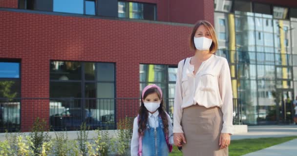 Glückliche kaukasische Familie, die in Masken im Freien steht. Schöne Mutter in Maske, die gut gelaunt mit einem netten kleinen Schulmädchen in der Nähe der Schule auf der Straße steht. Zurück zur Schule. Quarantäne-Konzept — Stockvideo