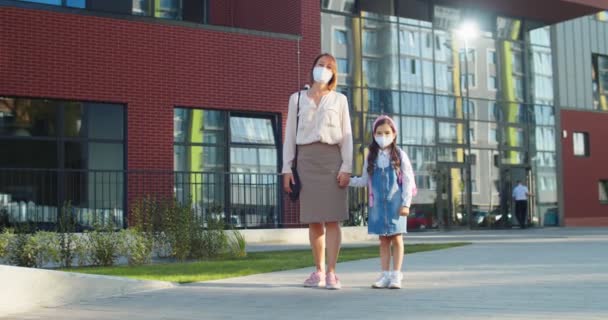 Güneşli bir günde maske takmış güzel beyaz bir bayan ve küçük tatlı bir kız öğrenci. Karantinada okul yakınlarında bir kızın elini tutan mutlu bir anne. Okula dönelim. Konsept çalış — Stok video