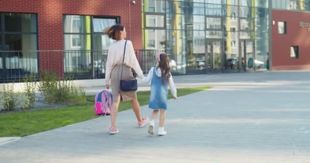 Küçük kızıyla birlikte okula yürüyen ve güneşli bir günde konuşan güzel beyaz bir kadının arkası. Anne kızı okula götürüyor ve elinde pembe sırt çantası eğitim konseptiyle konuşuyor. — Stok video
