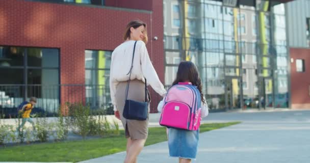 Pohled zezadu na krásnou bělošskou matku, jak se ráno prochází s malou školačkou do školy. Šťastná žena vede veselou dceru s růžovým batohem do tříd a mluví. Studijní koncept — Stock video