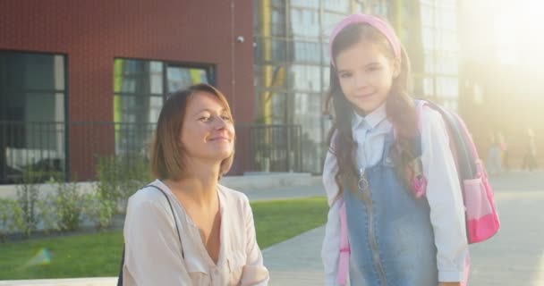 Retrato de mãe e filha caucasiana feliz sorrindo para a câmera perto da escola ao ar livre no dia ensolarado. Mulher alegre bonita com colegial bonitinho no pátio da escola após as aulas. Conceito de educação — Vídeo de Stock