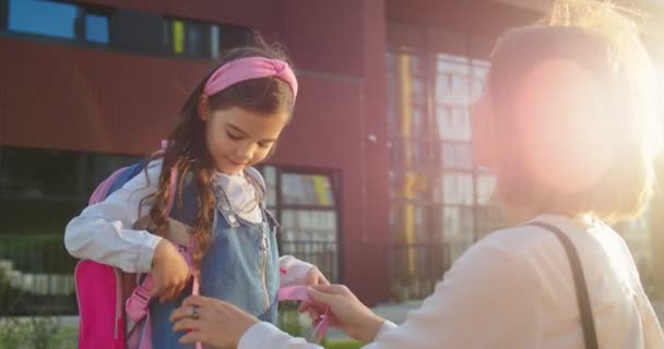 Närbild porträtt av vacker kvinna fastställande kläder på söt liten skolflicka utomhus. Sidovy över kaukasisk lycklig mor med dotter elev nära skolan på solig morgon. Tillbaka till skolkonceptet — Stockvideo