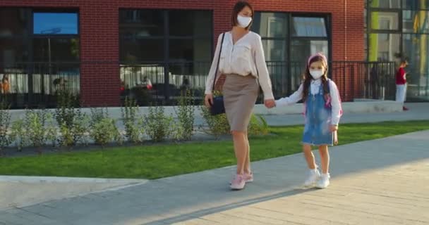 Kavkazská matka a roztomilá dcera žákyně v maskách kráčející venku za slunečného dne po vyučování. Krásná žena chodí s malou školačkou ze školy v karanténě a mluví. Koncept vzdělávání — Stock video