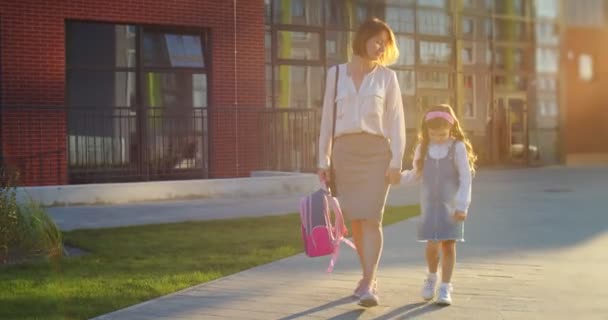 Kaukázusi anya és aranyos szomorú lánya sétál az iskolából a szabadtéri órák után a napsütéses napon. Csinos nő kis feldúlt iskolás lánnyal sétál az utcán a városban. A gyermeknevelés fogalma — Stock videók