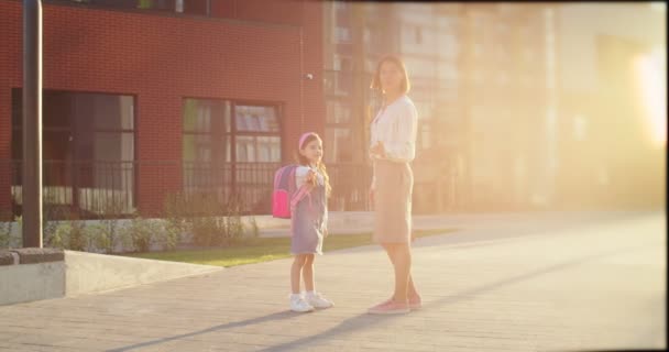Beyaz tenli güzel bir kadın, güneşli bir günde okulun yanında sevimli küçük bir çocukla konuşuyor. Küçük neşeli kız annesiyle sohbet ediyor ve dışarıda dururken bir şeyler gösteriyor. Öğrenci konsepti — Stok video