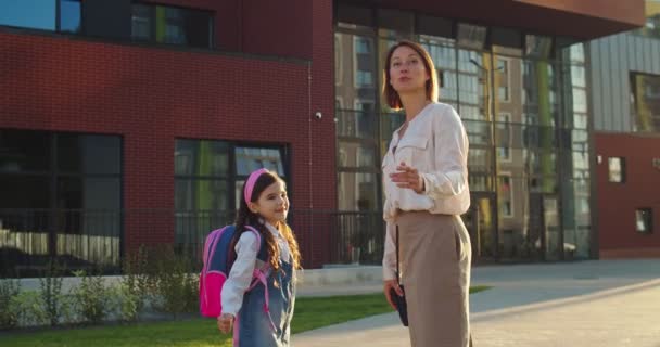 Красивая белая родительница разговаривает с милой школьницей возле школы в солнечный день. Маленькая счастливая девочка разговаривает с матерью и показывает что-то стоящее на улице. Концепция школы — стоковое видео