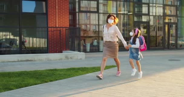 Kaukázusi boldog anya és aranyos kis iskolás lány maszkban sétál ki a szabadba a napsütéses napon az iskolából és ugrál. Elég vidám nő, kisgyerekkel a városban szaladgál és mosolyog. Karantén elve — Stock videók