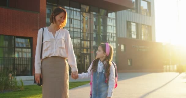 Кавказская веселая женщина держит за руку маленькую дочь и стоит рядом со школой на улице. Счастливая школьница держит мать за руку на солнце и улыбается в камеру. Снова в школу — стоковое видео