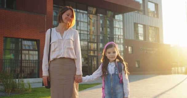 Kavkazský radostný ženský rodič držící malou dcerku za ruku a stojící u školy venku ve městě. Šťastná školačka s růžovým batohem stojí s matkou a usmívá se do kamery. Koncept vzdělávání — Stock video