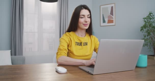 Vrolijk mooi blank meisje in gele trui opent laptop en het zoeken naar internet thuis. Vrij gelukkige jonge vrouw typen op de computer terwijl zitten aan tafel aan de binnenkant. Vrijetijdsconcept — Stockvideo