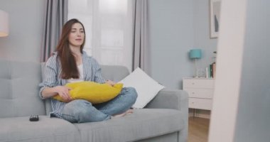 Güzel neşeli kız evde dinlenirken ve televizyon izlerken gülüyor. Oturma odasındaki gri kanepede oturan ve içeride televizyon izleyen güzel, beyaz, neşeli genç bir kadın. Boş zaman konsepti