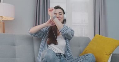 Evde kulaklıkla müzik dinlerken gülümseyen ve dans eden neşeli güzel bir kadın. Güzel neşeli kız oturma odasında kanepede otururken şarkı dinliyor ve komik hareketler yapıyor. Eğlenceli konsept