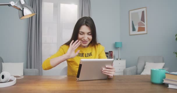 Alegre hermosa mujer joven caucásica agitando la mano y hablando en videollamada en la tableta en la habitación en casa. Feliz chica bonita sonriendo mientras tiene una conversación de vídeo en el interior del dispositivo. Concepto de ocio — Vídeos de Stock