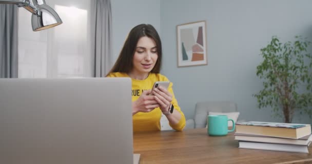 Vreugdevolle blanke mooie meisje sms 'en op de smartphone en glimlachen terwijl zitten aan het bureau in de kamer in huis. Mooie gelukkige jonge vrouw typen en tikken op mobiele telefoon terwijl ze binnen rust. Beginbegrip — Stockvideo