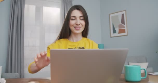 Vrolijk mooi meisje opent computer en typen tijdens het werken thuis in goed humeur. Mooie blanke vrolijke jonge vrouw browsen op laptop terwijl zitten aan tafel in de kamer. Freelancer-concept — Stockvideo