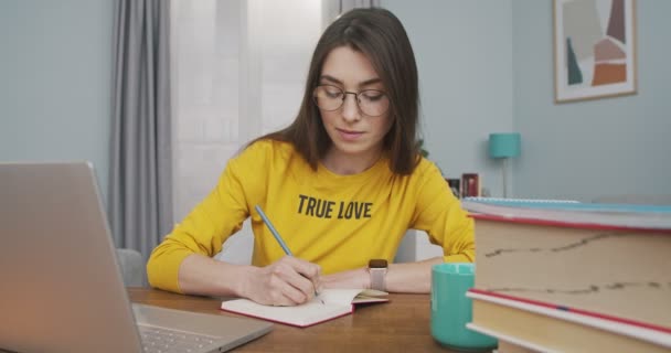 Portret młodej, ładnej białej kobiety w okularach, patrzącej na ekran laptopa i spisującej plan w domu. Piękna dziewczyna robi notatki do notatnika siedząc przy stole w pokoju Koncepcja badania — Wideo stockowe