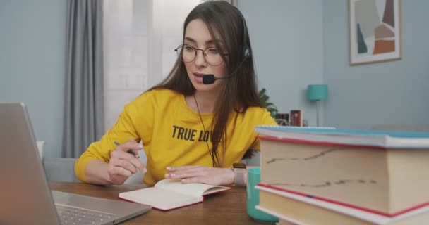 Porträtt av vacker vit kvinna i glasögon tittar på bärbar skärm och skriva ner i planerare hemma medan du pratar i headset. Vacker flicka att göra anteckningar i anteckningsbok när du sitter vid bordet — Stockvideo