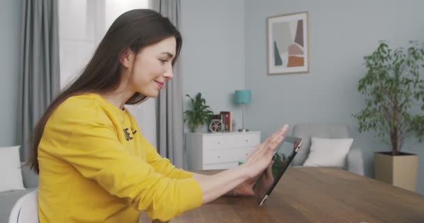 Zijaanzicht van vrij vrolijke jonge vrouw scrollen en surfen op tablet, terwijl zitten aan het bureau in de kamer thuis. Portret van een mooi blank gelukkig meisje dat binnen op het apparaat tikt. Rust begrip — Stockvideo