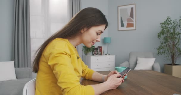 Side view van Kaukasische vrolijke mooie jonge meisje sms 'en op de smartphone, terwijl zitten aan het bureau in de kamer thuis. Portret van een mooie vrolijke vrouw die binnen typt en afluistert op een mobieltje. Vrijetijdsconcept — Stockvideo