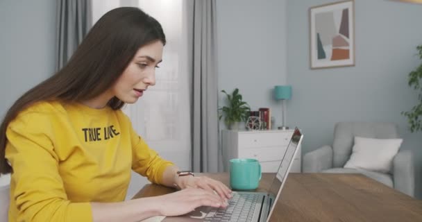 Närbild porträtt av koncentrerad söt flicka skriva på laptop när du sitter vid skrivbordet i mysiga rum hemma. Sidovy över vackra unga kvinna sms:ar på datorn inomhus. Frilansbegreppet — Stockvideo